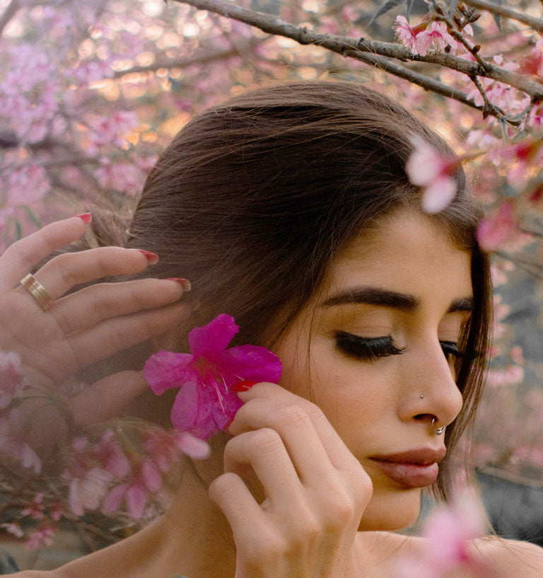 Woman with dark beautiful thick hair putting flower behind ear and surrounded with pink flowers gazing down. Thick lustrous healthy hair with Neelibringadi Nourishing and Moisturizing Hair Growth Oil from Vedic Tiger. 